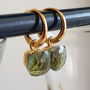 Puede incluir: Pendientes de aro de oro con colgantes de piedras preciosas verdes. Las piedras preciosas son facetadas y tienen una apariencia ligeramente translúcida.