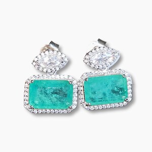 Puede incluir: Un par de pendientes de plata con piedras preciosas verdes vibrantes con talla esmeralda. Cada pendiente tiene una piedra rectangular con talla esmeralda rodeada por un halo de pequeños cristales transparentes. La parte superior de cada pendiente tiene un grupo de cristales transparentes en forma de marquesa.