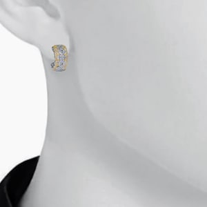 May include: A close-up of a single earring. The earring is a small, curved hoop with a textured surface. The top portion is gold-colored, while the rest is silver-colored. The earring is worn on a white surface.