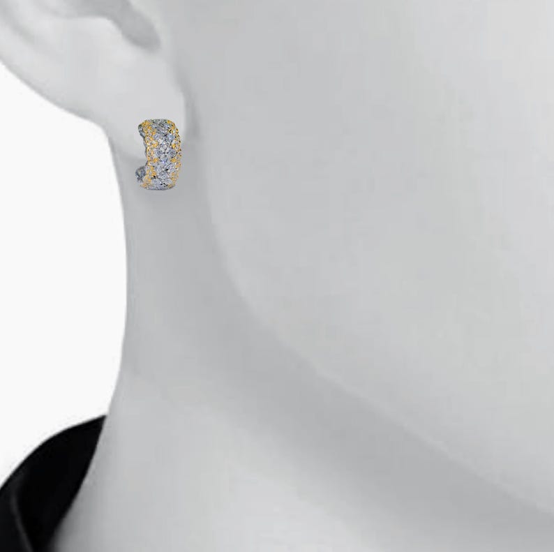May include: A close-up of a single earring. The earring is a small, curved hoop with a textured surface. The top portion is gold-colored, while the rest is silver-colored. The earring is worn on a white surface.