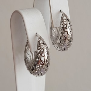 May include: A pair of silver hoop earrings with a delicate, intricate design. The earrings are made of metal and have a smooth, polished finish.