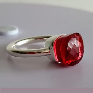 Puede incluir: Un anillo de plata con una gran gema roja facetada. La gema, de forma cuadrada, captura la luz, mostrando sus m&uacute;ltiples facetas. La banda es de plata pulida. El anillo se encuentra sobre un fondo claro.