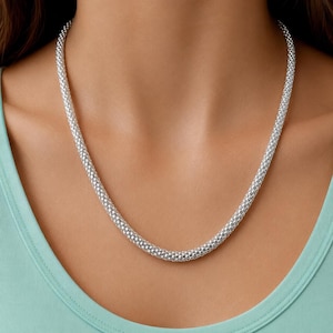 Può includere: Una collana a catena popcorn color argento caratterizzata da un design a maglie arrotondate e testurizzate con finitura metallica. La catena poggia sul collo, mostrando un motivo coerente e ripetitivo di piccole maglie sferiche collegate.