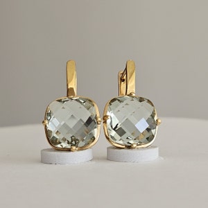 Op de afbeelding: Een paar goudkleurige oorbellen met vierkante, lichtgroene edelstenen. De edelstenen zijn facetgeslepen en de oorbellen hebben een klapsluiting. De oorbellen worden gepresenteerd op kleine, witte cilindrische standaards.