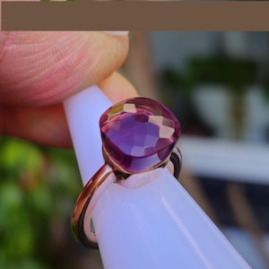 Puede incluir: Un anillo de oro rosa con una gran gema p&uacute;rpura facetada. La gema, de talla coj&iacute;n, captura la luz, mostrando varios tonos de p&uacute;rpura. El anillo se presenta sobre un fondo blanco, resaltando su dise&ntilde;o.