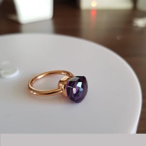 Puede incluir: Un anillo dorado con una gran gema p&uacute;rpura facetada. El anillo se muestra sobre una superficie blanca, resaltando el dise&ntilde;o y el color de la joya. La gema refleja la luz, agregando brillo al anillo.