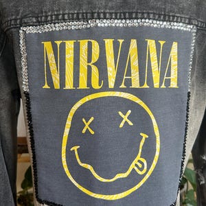 以下が含まれることがあります： チャコールグレーのデニムジャケット。背中には「NIRVANA」の黄色い文字と黄色いスマイリーフェイスのグラフィックが描かれた大きな長方形のパッチが付いています。パッチの上部は透明なラインストーン、側面と下部は黒いビーズで縁取られています。