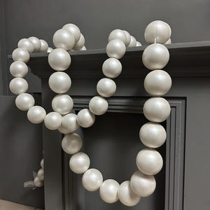 Pode incluir: Uma grinalda decorativa de grandes contas redondas brancas. As contas são enfiadas, criando um efeito fluido e drapeado. A grinalda é exibida contra um fundo cinza, adicionando um toque de elegância a qualquer espaço. Perfeita para decoração de casa.