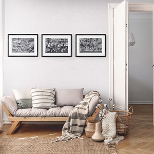 May include: A light gray futon sofa with a wooden frame and a striped throw blanket. The sofa is in a living room with a white wall, a window with white curtains, and a door leading to another room. There are three black framed pictures on the wall above the sofa.