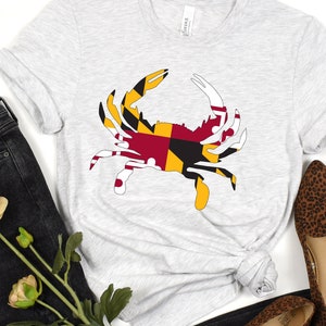 May include: A grey t-shirt with a white crab silhouette printed on it. The crab is filled with the colors of the Maryland state flag. The shirt is laid out on a white surface with a pair of brown leopard print shoes and black jeans.