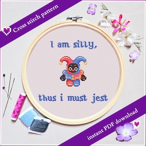 May include: A cross-stitch pattern in a wooden hoop features a jester design with the text "I am silly, thus I must jest." The design includes red, blue, and black colors. The image also shows embroidery supplies and flowers.