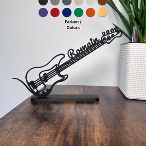 Può includere: Silhouette di chitarra in metallo nero con il nome "Romain" inciso sul manico. La chitarra è su un supporto e il nome è visibile. L'immagine include una tavolozza di colori di 12 colori diversi.
