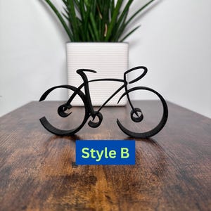 Könnte beinhalten: Eine schwarze Metall-Fahrrad-Skulptur im minimalistischen Stil. Die Skulptur steht auf einer Holzoberfläche und der Text "Style B" ist unterhalb der Skulptur sichtbar.