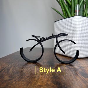 May include: Black metal bike-shaped ring holder with the name "Alexander" written on it. The holder is on a wooden surface. Style A.