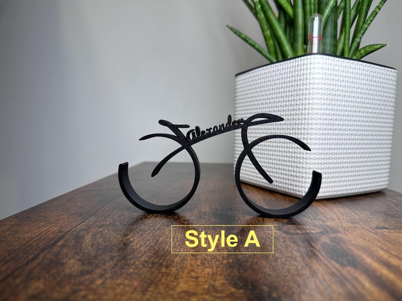 May include: Black metal bike-shaped ring holder with the name "Alexander" written on it. The holder is on a wooden surface. Style A.