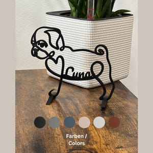 May include: Black metal silhouette of a pug dog with the name "Luna" written inside a heart. The silhouette is standing on a wooden surface. The image also shows a row of six colour swatches: black, grey, blue, beige, light blue, and brown.
