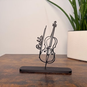 May include: Black metal violin sculpture with the name "Pablo" in script. The sculpture includes a violin and bow, mounted on a rectangular base. The sculpture is displayed on a wooden surface.