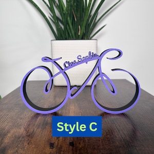 May include: Purple wooden bicycle silhouette with the names "Emma Sophia" written on it. The bicycle is on a wooden surface. The text "Style C" is below the bicycle.