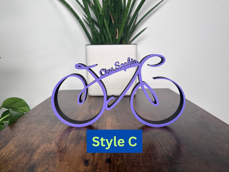 May include: Purple wooden bicycle silhouette with the names "Emma Sophia" written on it. The bicycle is on a wooden surface. The text "Style C" is below the bicycle.
