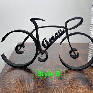 May include: Black wooden bicycle silhouette with the name "Aryan" written in cursive on the frame. The bicycle is on a wooden surface with the text "Style B" below it.