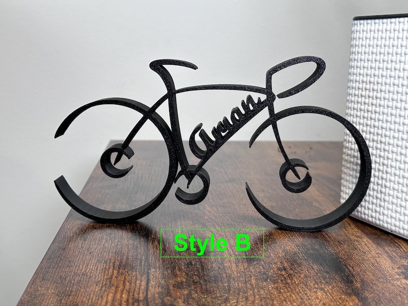 May include: Black wooden bicycle silhouette with the name "Aryan" written in cursive on the frame. The bicycle is on a wooden surface with the text "Style B" below it.