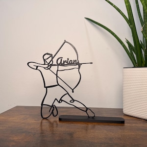 Könnte beinhalten: Schwarze Metall-Bogenschützen-Silhouette mit dem Namen "Arian" auf dem Bogen. Der Schütze befindet sich in einer Schusspose, montiert auf einem schwarzen rechteckigen Sockel. Eine Topfpflanze in einem weißen strukturierten Topf befindet sich im Hintergrund.