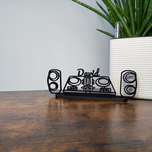 Könnte beinhalten: Schwarzer, lasergeschnittener DJ-Schreibtisch-Organizer mit dem Namen "David" in Schreibschrift. Der Organizer zeigt Lautsprecher- und Mischpult-Designs. Ein weißer, strukturierter Pflanztopf mit einer grünen Pflanze befindet sich im Hintergrund.