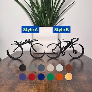 May include: Two black metal bike sculptures, one in a simple design and the other with more detail, with the words "Sarah" and "Joshua" incorporated into the design. The sculptures are on a wooden surface with a variety of color options displayed below.