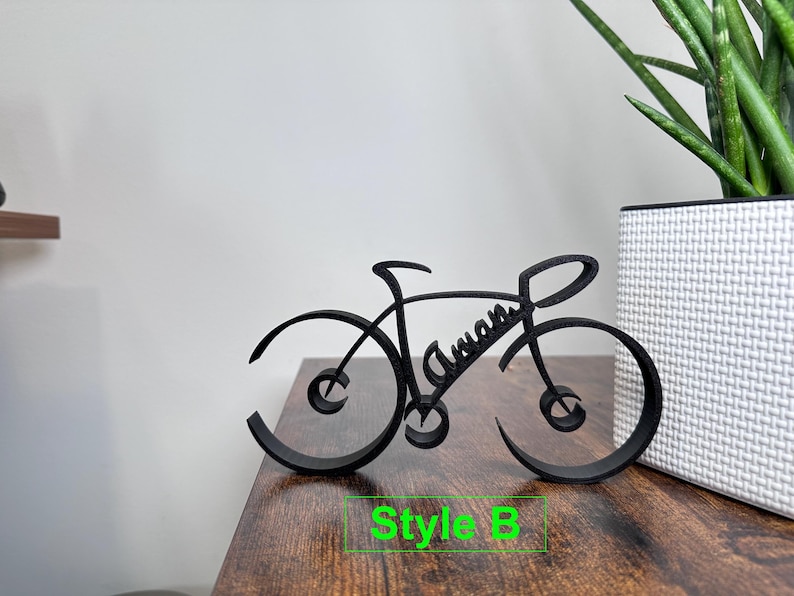 May include: Black metal bicycle silhouette with the name "Aman" written on the frame. The bicycle is on a wooden surface with the text "Style B" below it.