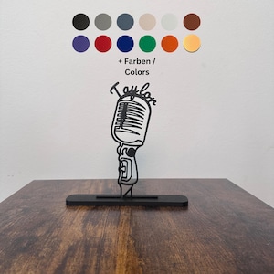Puede incluir: Decoración de metal negro con forma de micrófono con el nombre "Taylor" en escritura cursiva. La decoración está sobre una base negra, con una paleta de colores variados encima. El micrófono mide aproximadamente 15 cm de alto.