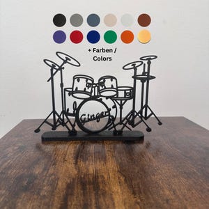 May include: A black, laser-cut drum kit silhouette with cymbals, drums, and stands. The drum kit has the name "Ginger" on the bass drum. A colour palette of various colours is displayed above the drum kit. The drum kit is on a wooden surface.