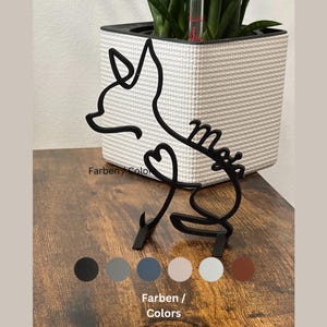 Könnte beinhalten: Schwarze Metall-Silhouette eines Fuchses mit einem Herzen und dem Wort "Mia" in Kursivschrift. Die Silhouette befindet sich auf einer Holzoberfläche mit einem weißen Topf und einer grünen Pflanze im Hintergrund. Das Bild zeigt Farboptionen für die Silhouette: Schwarz, Grau, Blau, Beige, Hellblau und Braun.