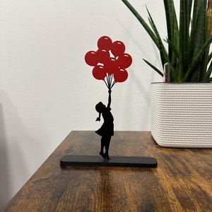 Puede incluir: Silueta de metal negro de una niña sosteniendo un ramo de globos rojos. La silueta está sobre una base negra.