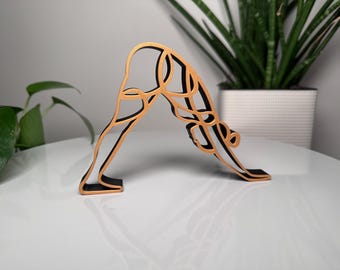 Könnte beinhalten: Eine Holzskulptur einer Yoga-Pose, dem herabschauenden Hund. Die Skulptur hat einen schwarzen Sockel und Kanten mit goldfarbenen Konturen. Die Yoga-Figur wird auf einer weißen Oberfläche mit einer grünen Pflanze im Hintergrund gezeigt.