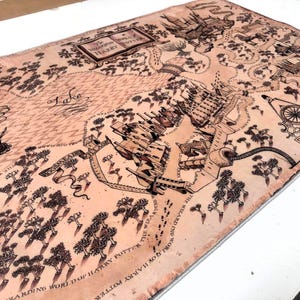 May include: A detailed map with a vintage aesthetic, featuring intricate illustrations of a fantasy landscape. The map is predominantly tan with black ink detailing, including text and various symbols. The text reads "WIZARDING WORLD OF HARRY POTTER".