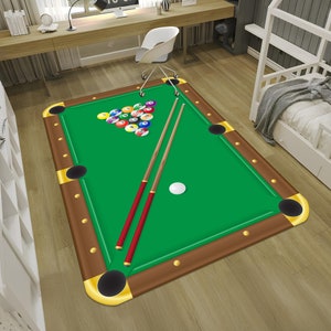 May include: A green felt pool table rug with brown wooden rails and gold accents. The rug features a set of billiard balls in a triangle formation and two pool cues.
