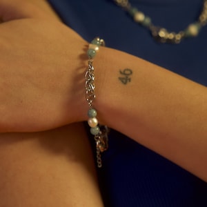 May include: A silver chain bracelet with light blue and white beads. The bracelet is on a person's wrist, and the number 46 is tattooed on their wrist.
