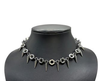 May include: Silver-toned choker necklace with a punk rock aesthetic. The necklace features a chain of hexagonal nuts and bolts, with evenly spaced, downward-facing, conical spikes. The necklace is displayed on a black mannequin.