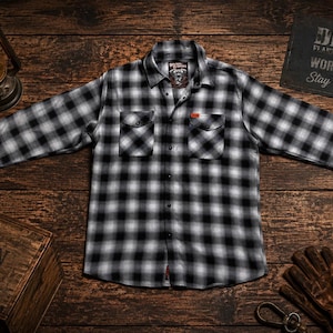 May include: Black and white plaid flannel shirt with two chest pockets and a red accent tag. The shirt is laid out on a wooden surface with a lantern, a wooden box, and a sign that reads "Work Hard. Stay Humble."