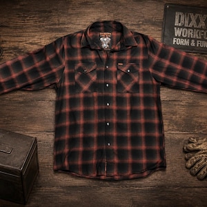 May include: A black and red plaid button-down shirt with two chest pockets. The shirt is displayed on a wooden surface, alongside a toolbox, work gloves, and a sign that reads "DIXON WORKFORCE FORM & FUNCTION". A lantern and chain are also visible.