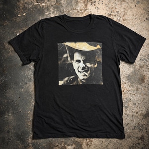 May include: A black short-sleeve t-shirt featuring a square graphic print on the chest. The print displays a high-contrast, sepia-toned photographic portrait of a person wearing a wide-brimmed hat with a cigarette held between their teeth.