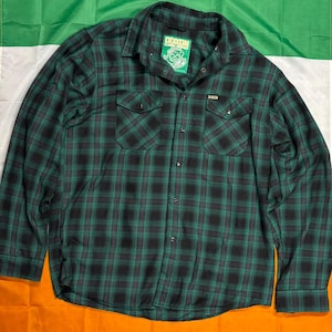 May include: A green and black plaid flannel shirt with two chest pockets and a button-down front. The shirt has a collar and long sleeves. The brand name "DIXON" is visible on a tag and on the shirt. The shirt is displayed on a green, white, and orange background.