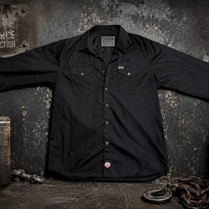 May include: Black button-down shirt with two chest pockets and a small tag. The shirt is spread out on a textured surface. The background includes a metal box, a barrel, and chains.