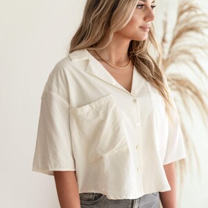 May include: A short-sleeved, off-white button-down shirt featuring a collared neckline and a large, square patch pocket on the front. The shirt has a relaxed, cropped fit and is made of a textured fabric, paired with grey denim jeans.