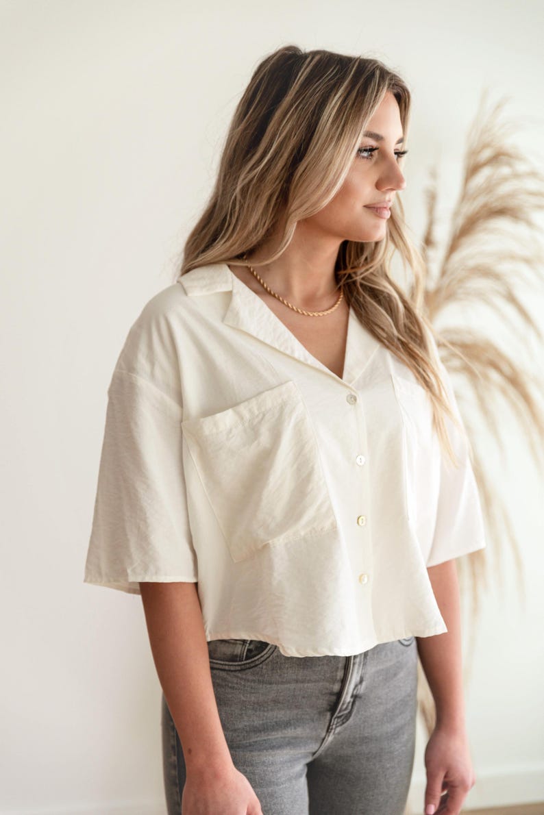 May include: A short-sleeved, off-white button-down shirt featuring a collared neckline and a large, square patch pocket on the front. The shirt has a relaxed, cropped fit and is made of a textured fabric, paired with grey denim jeans.