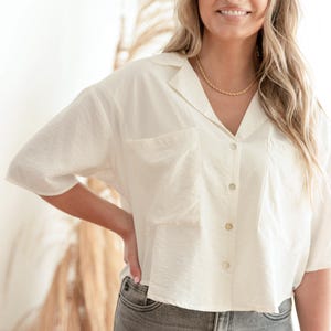 May include: A short-sleeved, off-white button-down shirt featuring a notched collar, two large front chest pockets, and a cropped hemline. The shirt is paired with grey denim jeans and a simple gold-toned chain necklace worn around the neck.