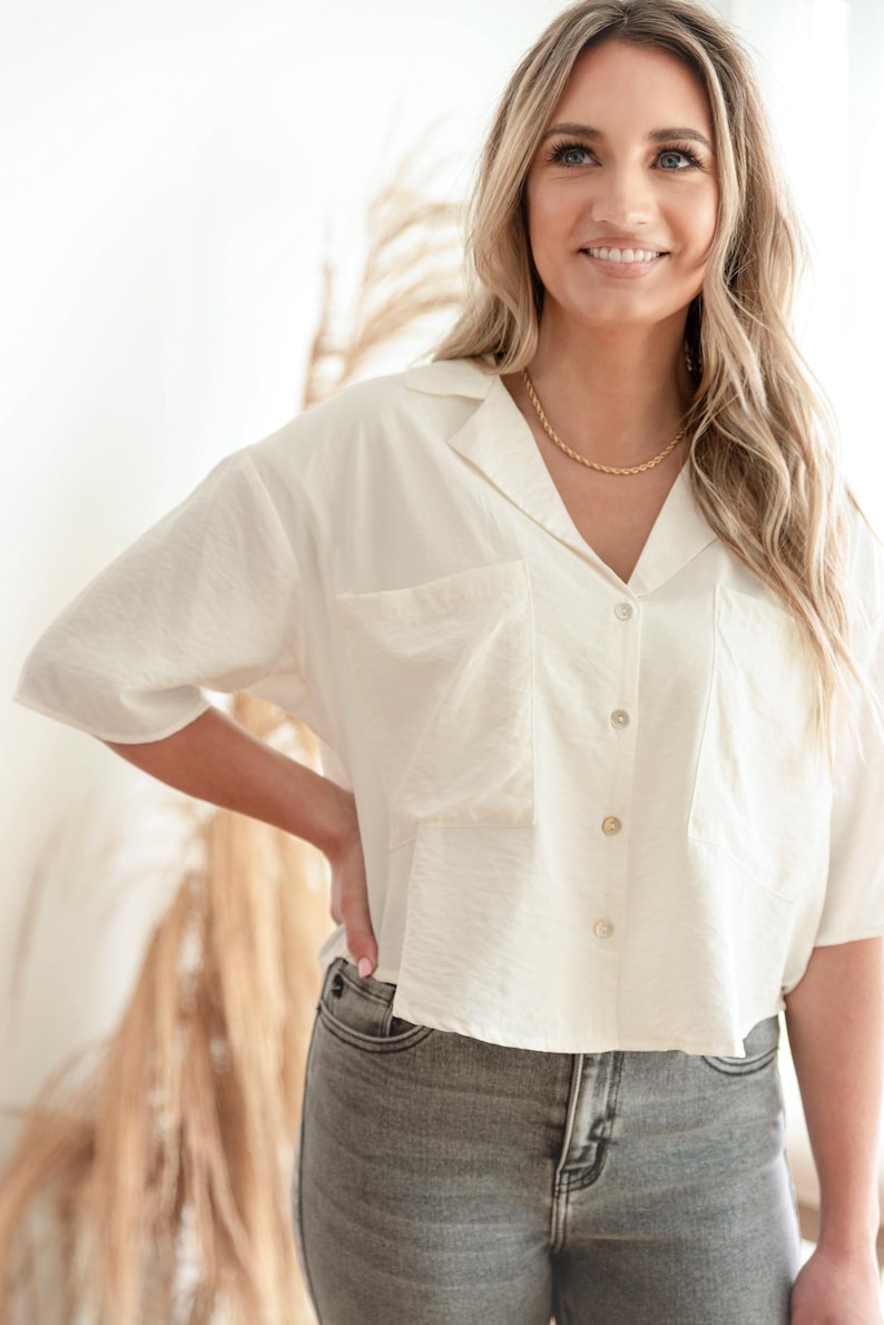 May include: A short-sleeved, off-white button-down shirt featuring a notched collar, two large front chest pockets, and a cropped hemline. The shirt is paired with grey denim jeans and a simple gold-toned chain necklace worn around the neck.