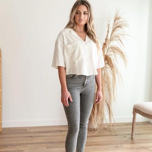 May include: A short-sleeved white button-down shirt with a collar and a single chest pocket, paired with grey skinny jeans and black slide sandals. The shirt has a relaxed, cropped fit, while the jeans feature a classic five-pocket design.