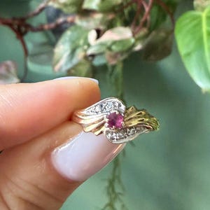 Puede incluir: Un anillo de oro con una piedra preciosa rosa de corte redondo en el centro, flanqueada por dos secciones de oro curvas y texturizadas. Cada sección de oro está acentuada con tres pequeñas piedras claras de corte redondo en fila.