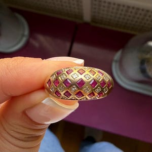 May include: A gold dome ring featuring a lattice pattern set with alternating square-cut red rubies and small round white diamonds. The band has a polished finish and a rounded profile, showcasing a repeating geometric design across the surface.